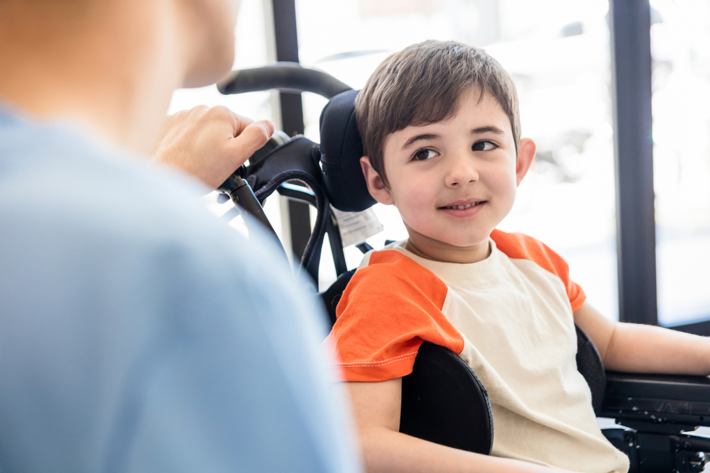 Beautiful little boy on wheelchair smiling and looking with admiration at his unrecongizable physical therapist at the clinic